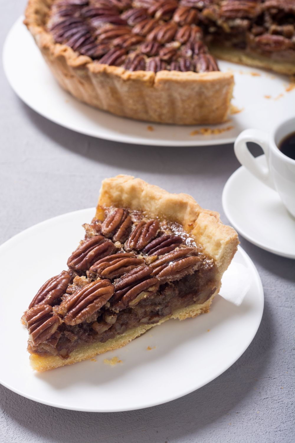 Homemade pecan pie on a white plate next to a cup of coffee